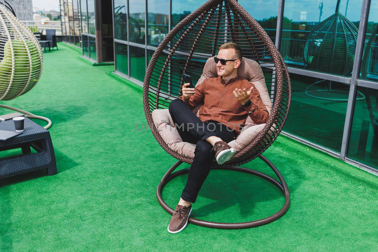 A cheerful young man in a shirt with a phone sits on the terrace of an office building after work, uses wireless internet, a positive male employee in glasses is talking on the phone
