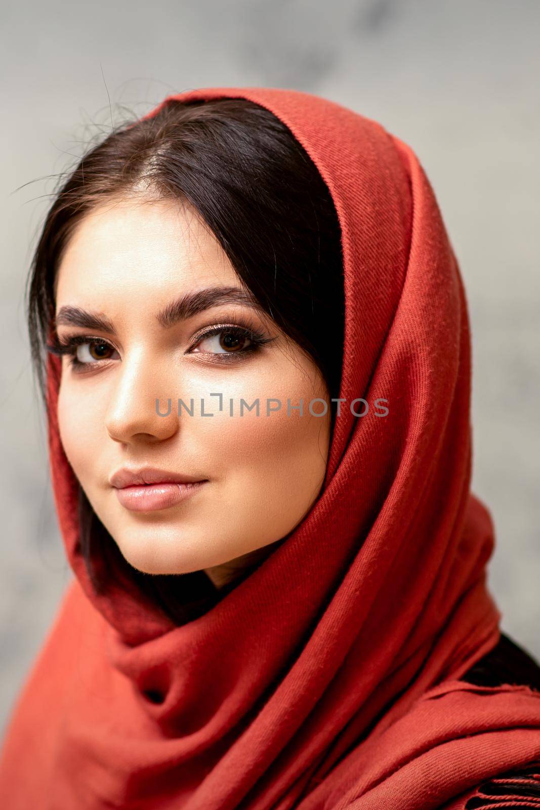 The fashionable young woman. Portrait of the beautiful female model with long hair and makeup in a red scarf. Beauty young caucasian woman on the background of a gray wall