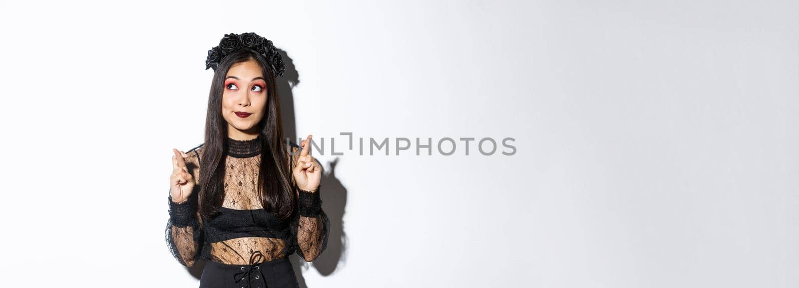 Hopeful cute asian woman in gothic lace dress and black wreath, looking upper left corner and cross fingers for good luck, making wish or praying, standing over white background by Benzoix