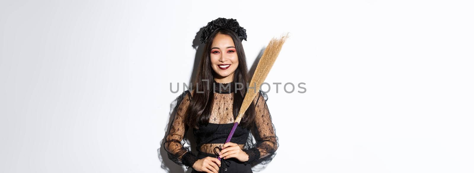 Portrait of smiling beautiful asian woman in witch costume holding broom and looking happy at camera, celebrating halloween, enjoying trick or treating, white background.