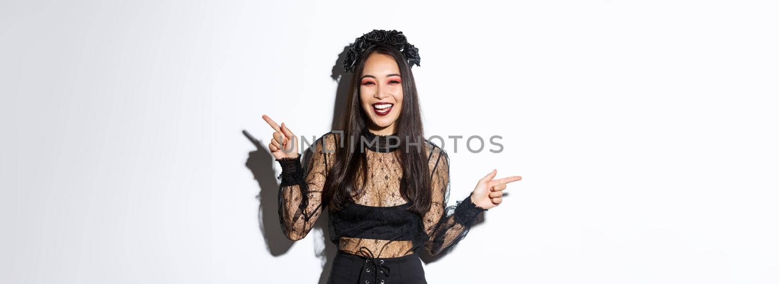 Beautiful happy young woman celebrating halloween in black lace dress, gothic style makeup, pointing fingers sideways, showing two banners or products, standing over white background.