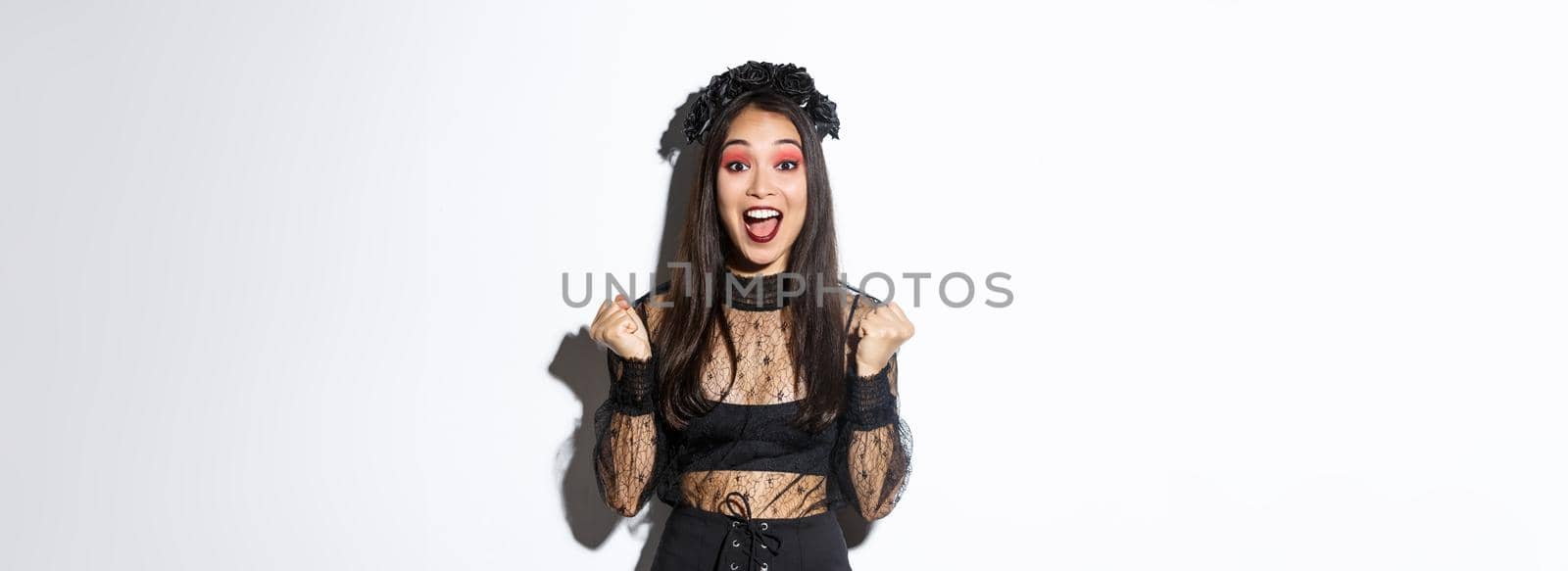 Image of happy asian gothic girl in halloween party dress, rejoicing over victory, triumphing and saying yes, standing over white background celebrating victory.