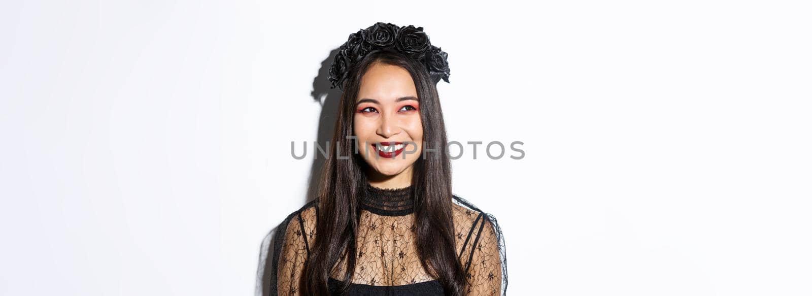 Close-up of beautiful elegant woman in witch costume and gothic makeup, smiling pleased while looking at upper left corner, celebrating halloween.