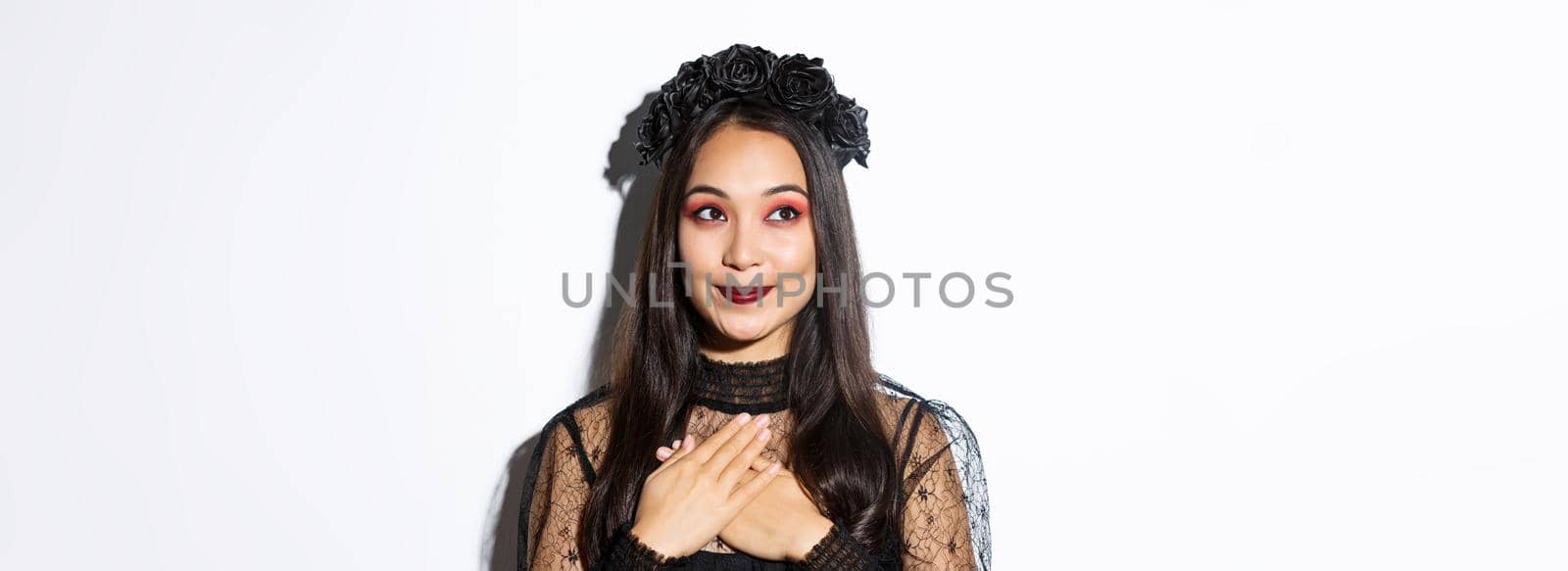 Close-up of nostalgic smiling asian woman looking at upper left corner dreamy, holding hands on heart, wearing gothic lace dress for halloween party.
