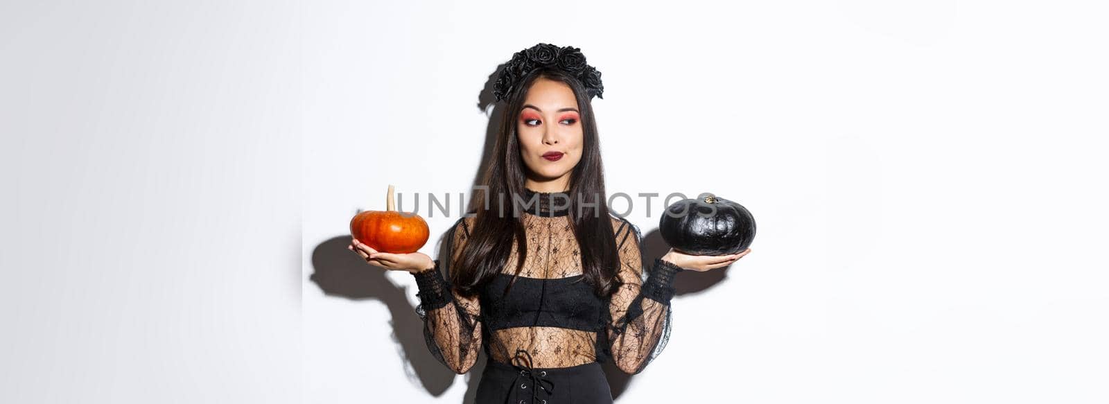 Thoughtful stylish woman in witch costume, picking pumpkin for halloween, making decision while standing over white background.