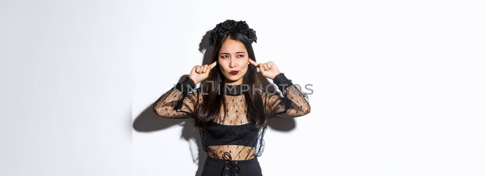 Annoyed and bothered asian stylish woman in halloween costume complaining on something loud, looking left displeased, shut ears with fingers, standing in gothic lace dress over white background.