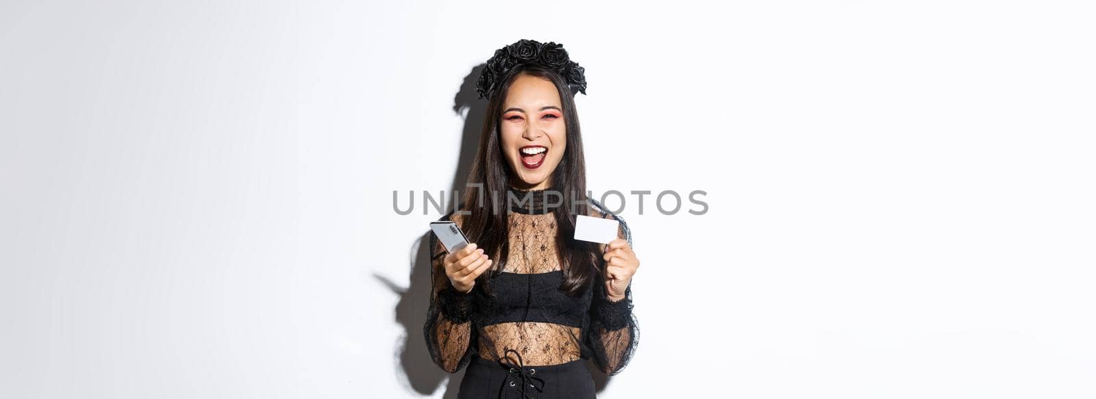 Happy laughing asian girl in halloween costume looking upbeat, holding smartphone and credit card, standing over white background by Benzoix