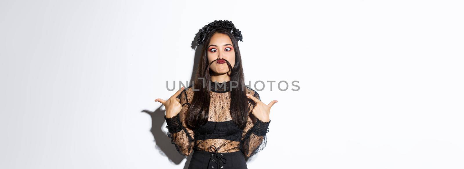 Portrait of funny and cute asian woman in witch costume making silly grimaces, pointing at herself, made moustache from hair strand.
