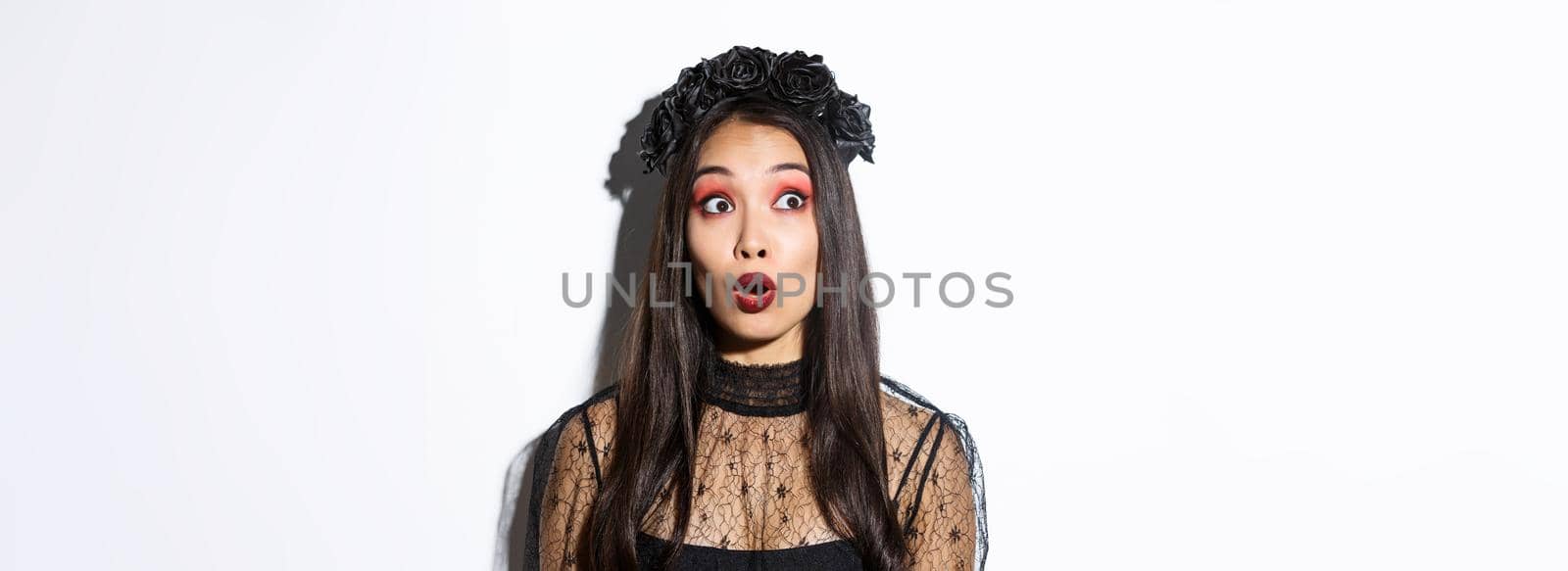 Close-up of surprised asian woman in witch costume looking at upper left corner and open mouth wondered, standing over white background.