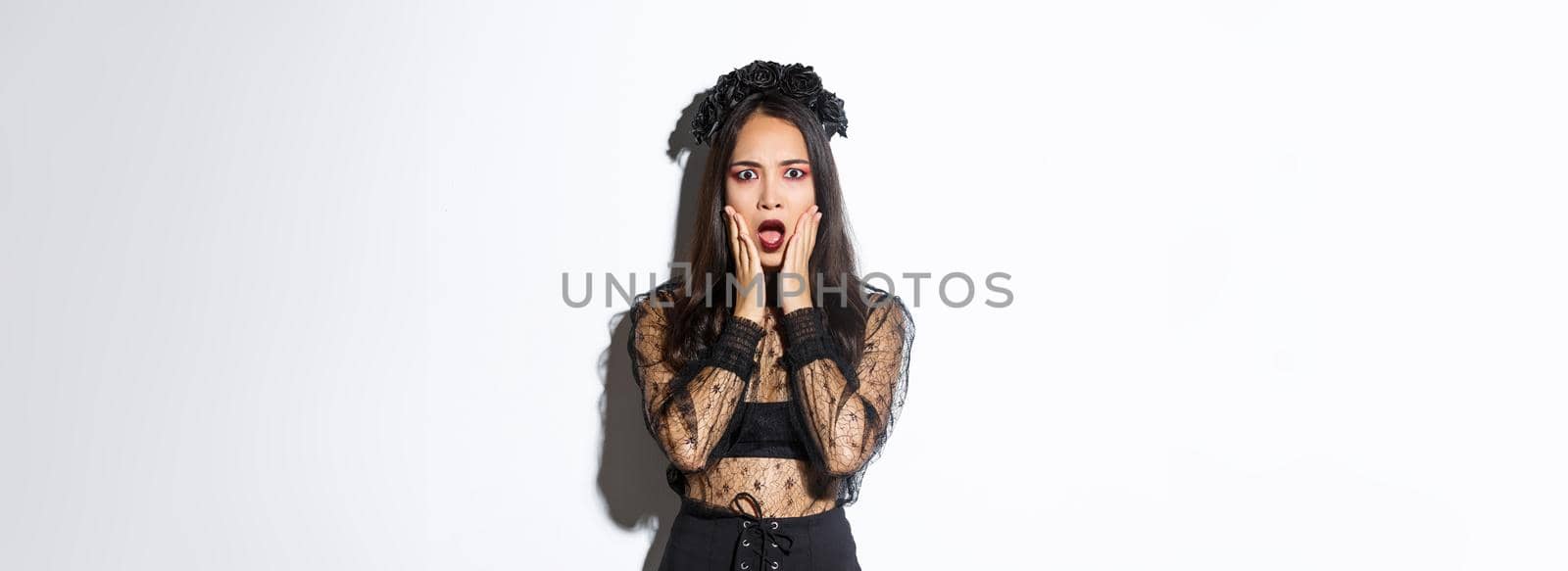 Shocked young asian woman wearing witch costume, looking concerned. Female in black dress and wreath looking like widow or magician gasping worried, standing white background.