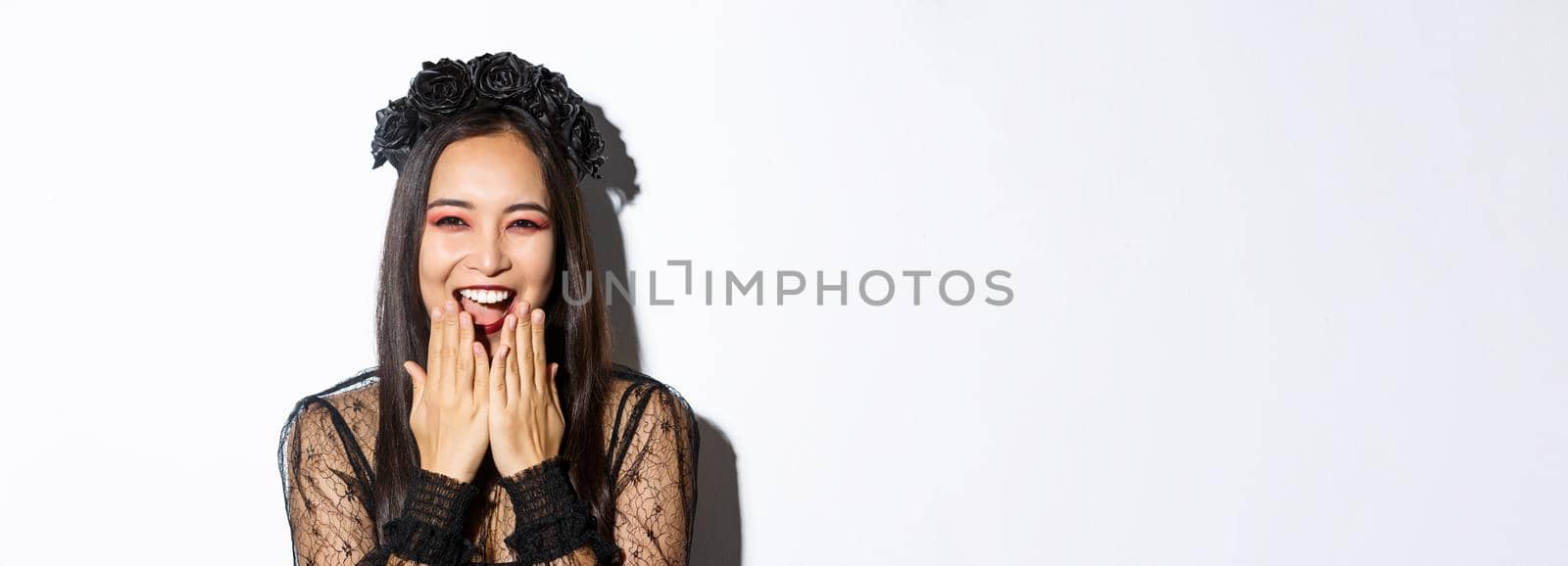 Close-up of happy asian woman celebrating halloween in witch costume and laughing, standing over white background by Benzoix
