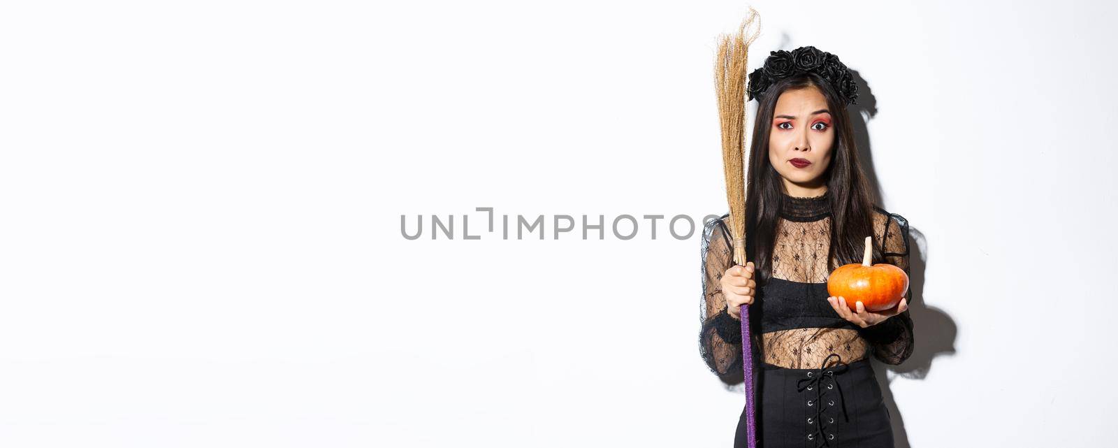 Worried and confused asian woman in witch costume looking nervous, holding broom and pumpkin, trick or treating on halloween, standing over white background.