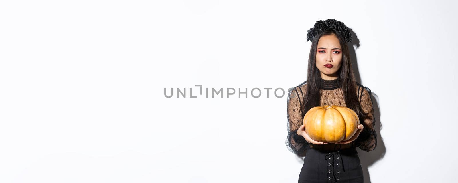 Image of grimacing asian woman in witch costume hate carve pumpkin for halloween, looking disappointed, standing over white background by Benzoix