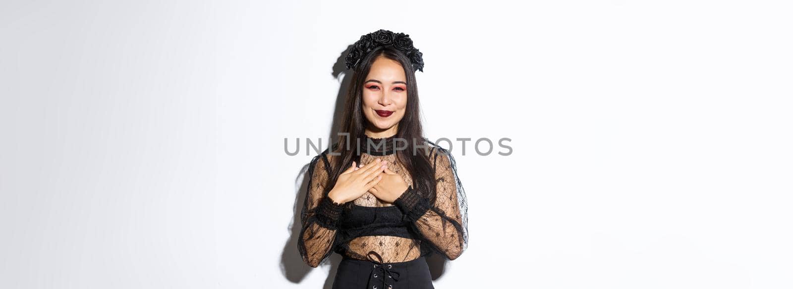 Pleased beautiful asian woman in witch costume smiling, holding hands on chest and looking grateful, express gratitude, standing in halloween outfit over white background by Benzoix