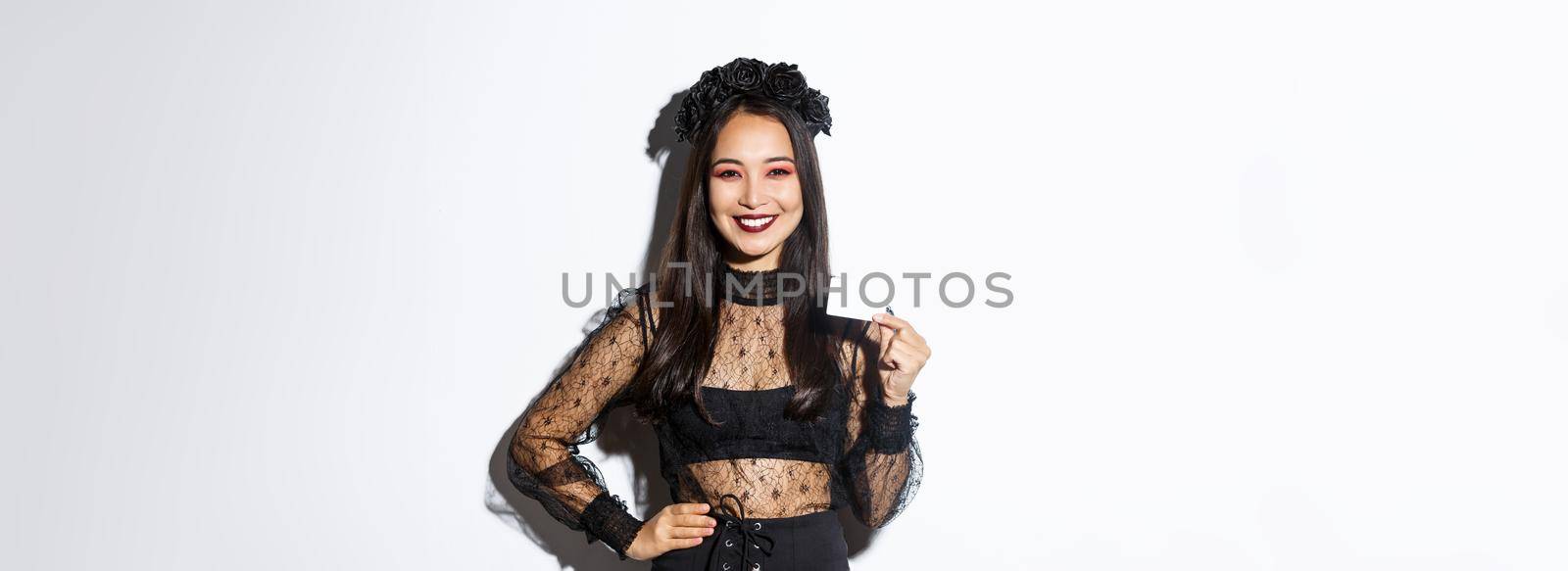 Image of beautiful asian woman in halloween costume, showing credit card and smiling, standing in gothic lace dress over white background.