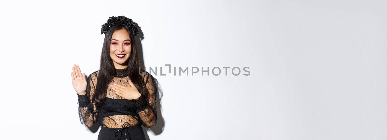 Image of cute sincere asian girl in halloween costume making promise, holding one hand on heart while swearing, being honest, standing over white background.