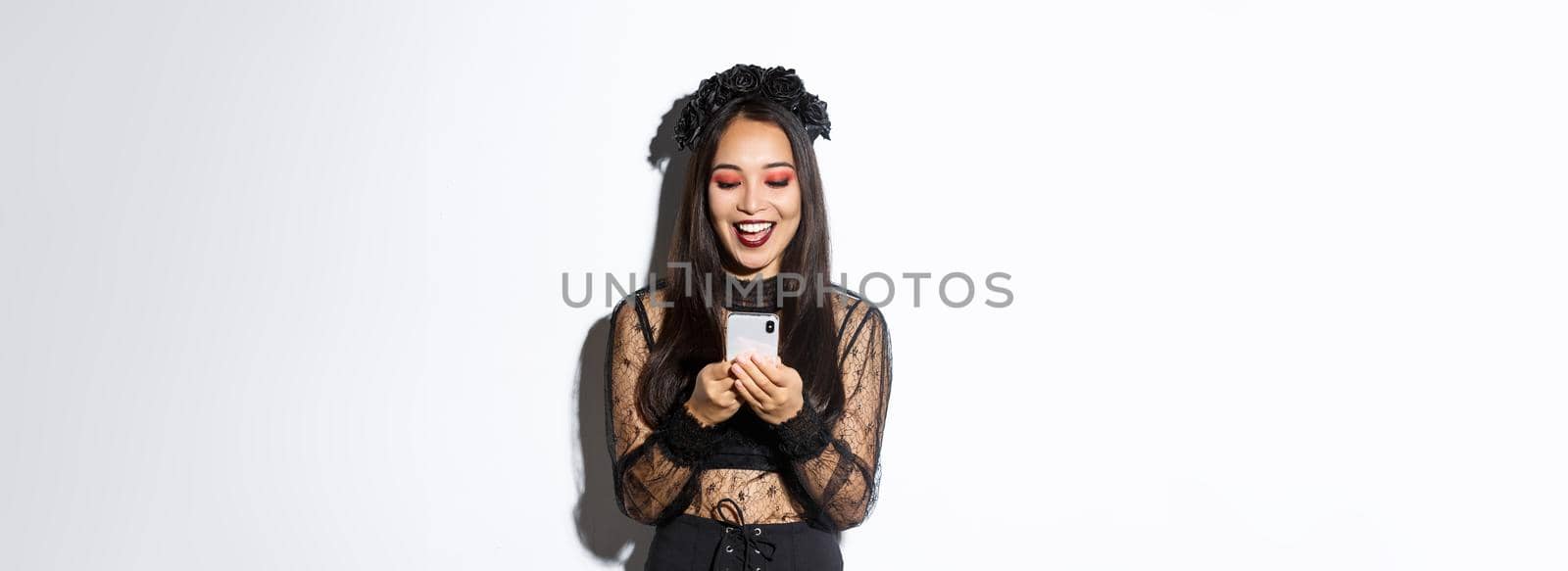 Excited smiling woman looking at mobile phone pleased, wearing gothic lace dress for halloween party, standing over white background.