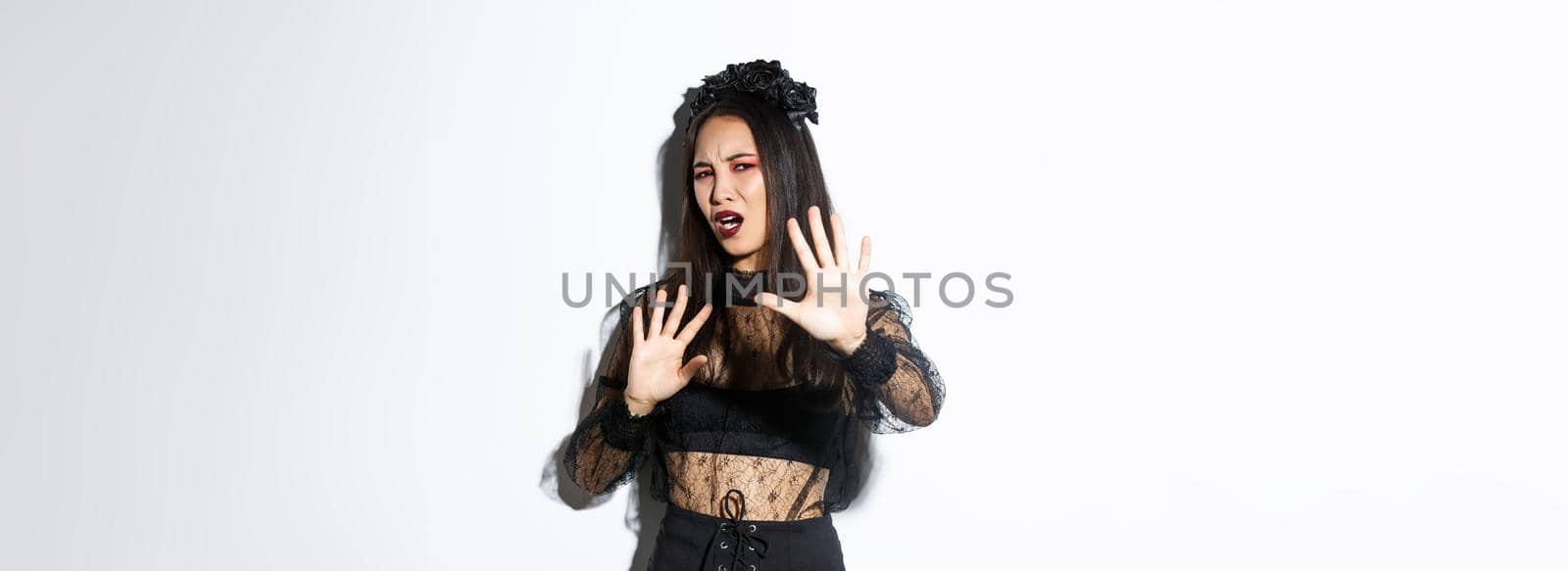 Image of bothered and annoyed asian woman in elegant gothic dress raising hands defensive, grimacing from camera flesh, asking to stop taking pictures of her, standing white background.