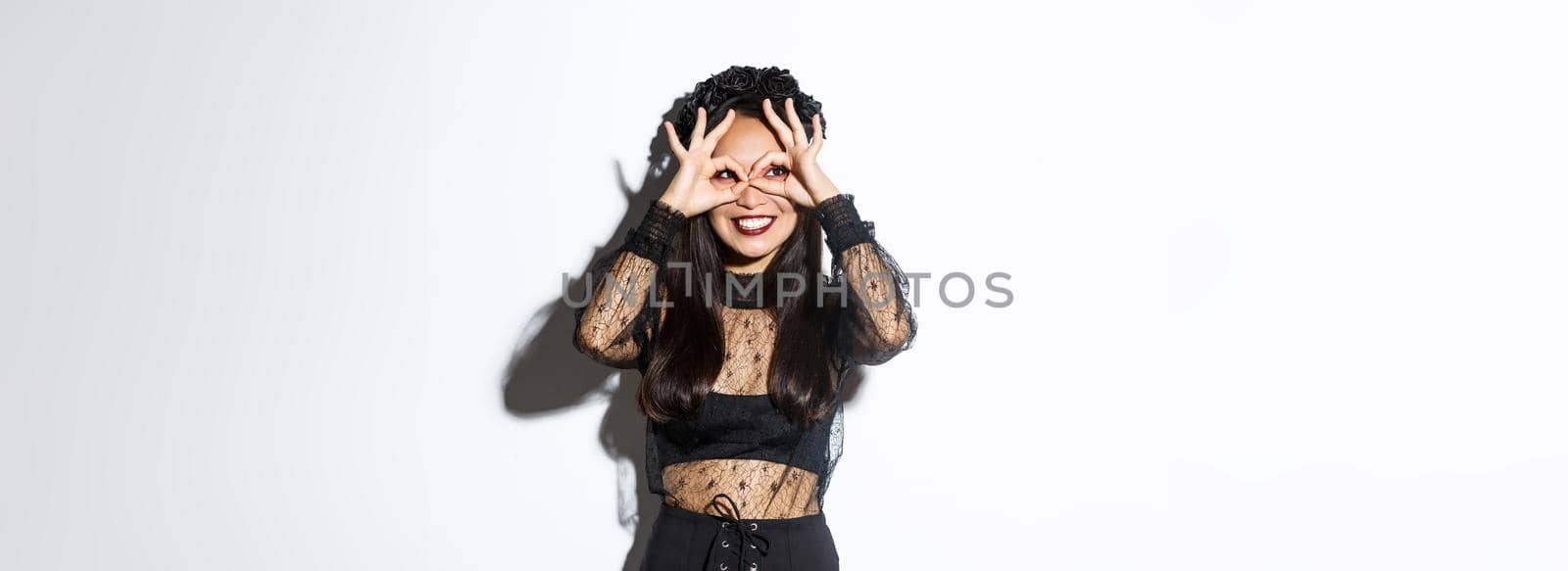 Portrait of beautiful smiling asian woman in halloween dress looking through fingers glasses at upper left corner, standing over white background by Benzoix