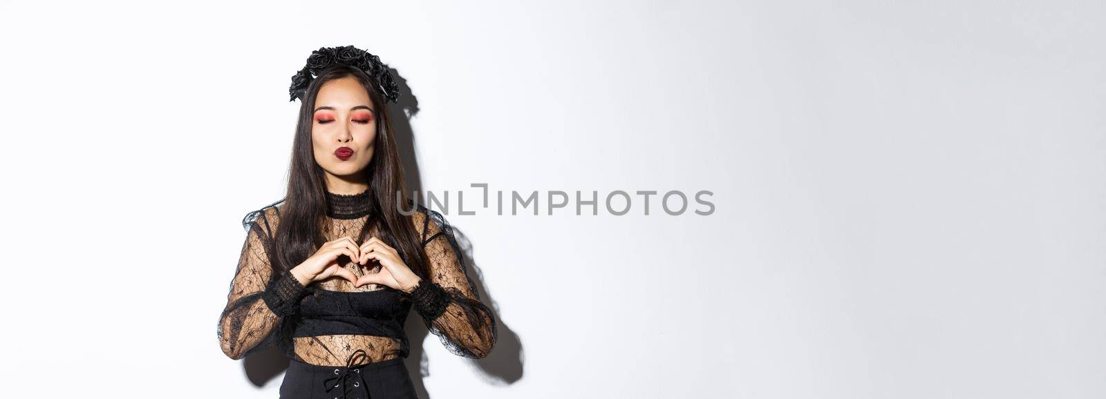 Image of beautiful asian gothic girl close eyes and pouting for kiss, showing heart gesture, express love or sympathy on halloween party, standing over white background by Benzoix