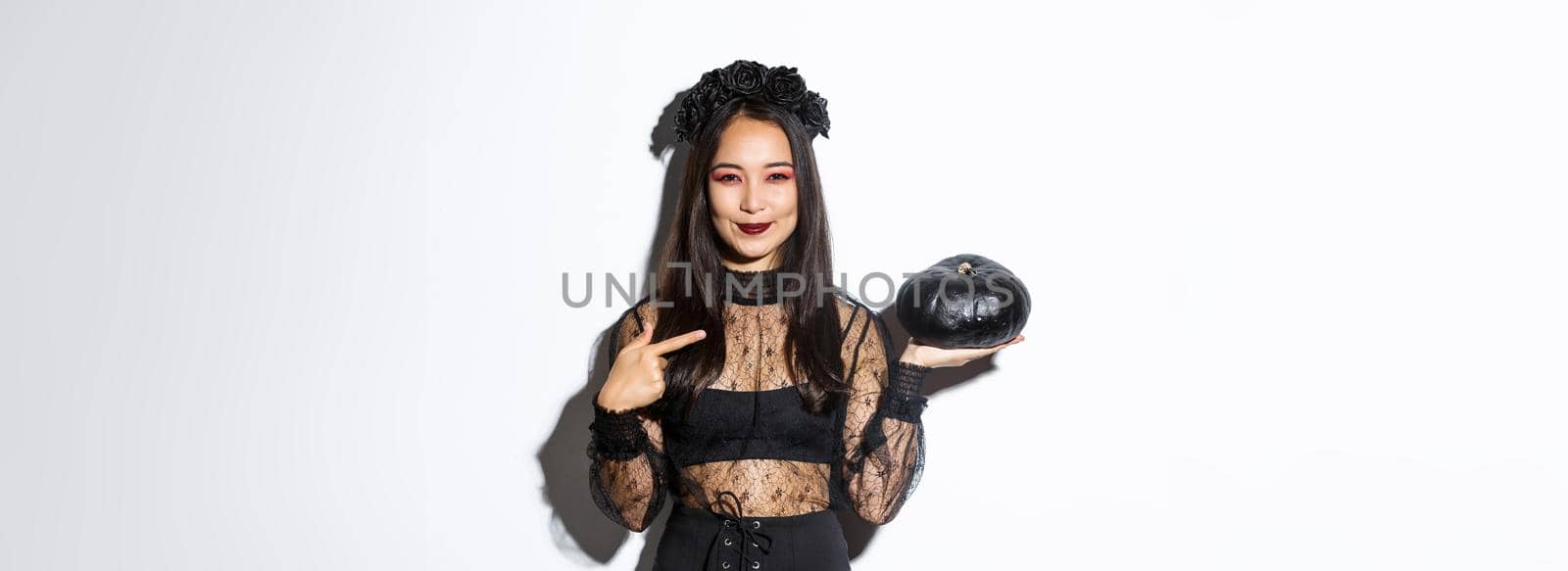 Image of beautiful asian woman in gothic lace dress and wreath pointing finger at black pumpkin, celebrating halloween, wearing witch costume, standing over white background.