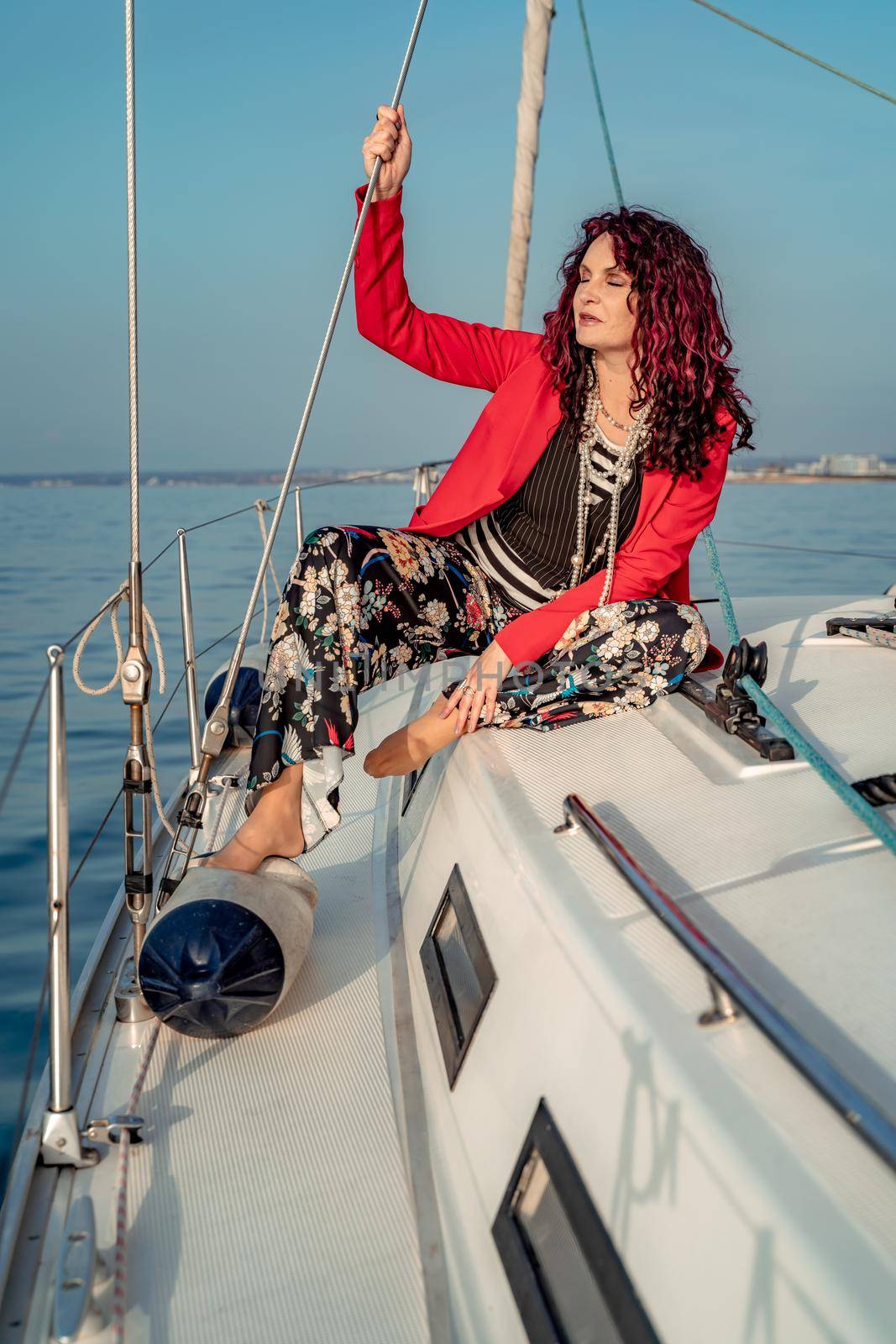 A woman sits on the bow of a yacht on a sunny summer day, the breeze develops her hair, a beautiful sea is in the background.