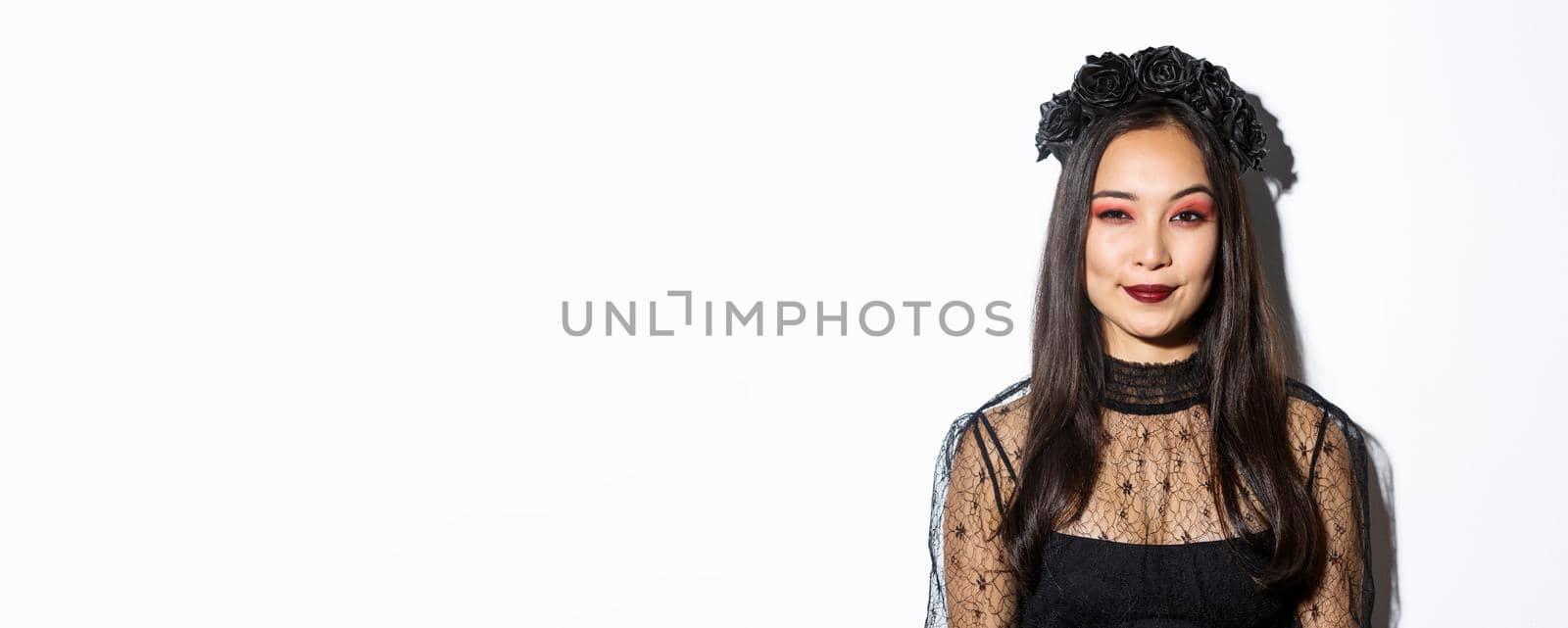 Image of smiling devious young witch looking at camera with cunning smirk, standing over white background. Woman in witch costume celebrating halloween.