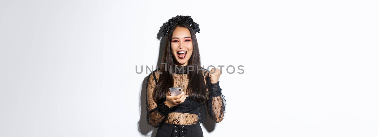 Image of successful happy asian woman celebrating good news, fist pump in rejoice and holding smartphone, wearing halloween costume, standing over white background.
