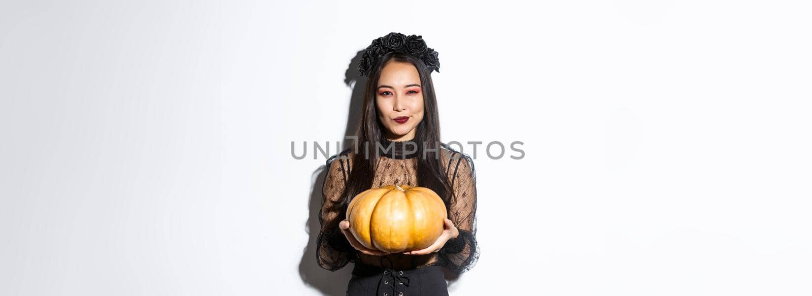 Image of cunning asian woman in black dress, impersonating evil witch on halloween, holding big pumpkin, standing over white background by Benzoix