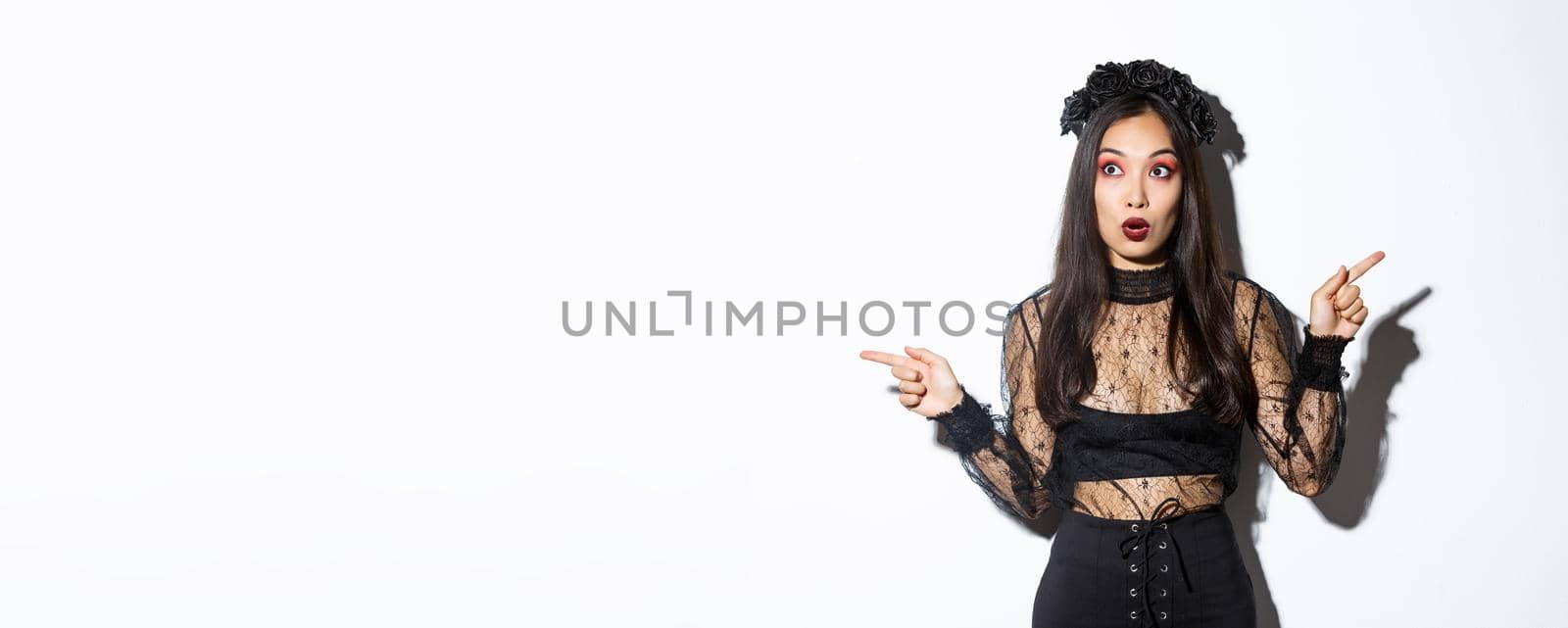 Amazed and startled woman in witch costume looking impressed, pointing fingers sideways but stare left, open mouth wondered, standing in black lace dress and wreath, white background.