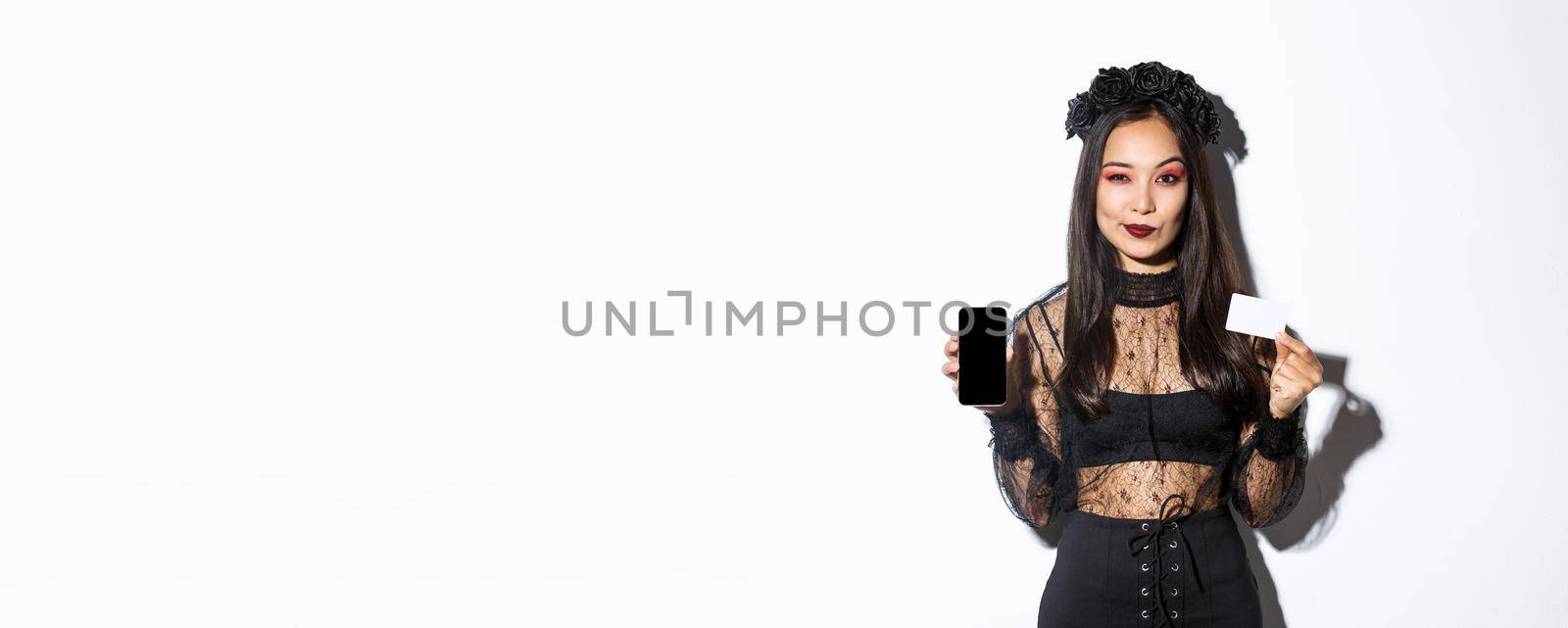 Sassy elegant young witch in gothic lace dress and black wreath showing you credit card and mobile phone screen, standing over white background.