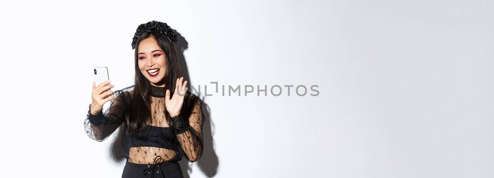 Portrait of beautiful and stylish asian woman in gothic lace dress saying hi, waving hand at smartphone camera during video call, standing over white background.