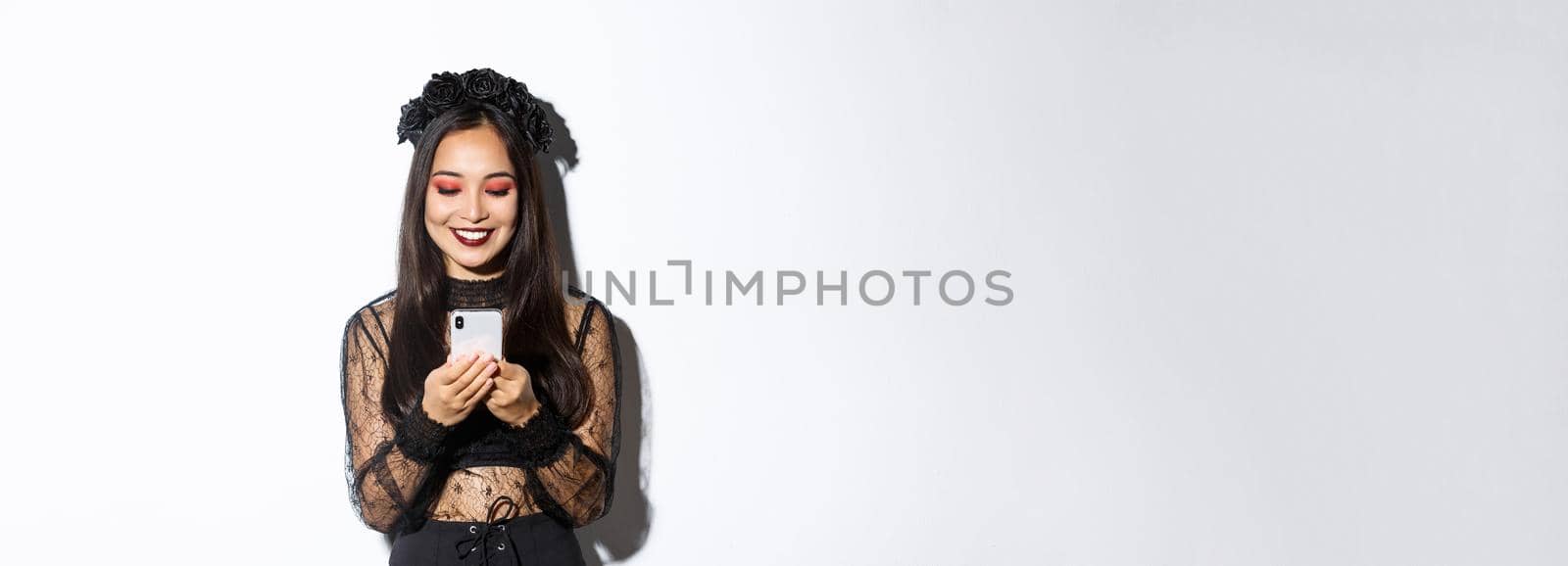Image of beautiful asian woman in gothic lace dress impersonating witch on halloween party, standing with smartphone and smiling over white background.