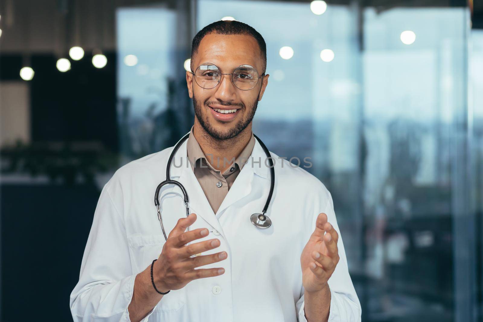 Close-up photo of hispanic doctor smiling and looking at web camera talking to colleagues and patient on video call, man in medical coat and stethoscope working inside clinic office.