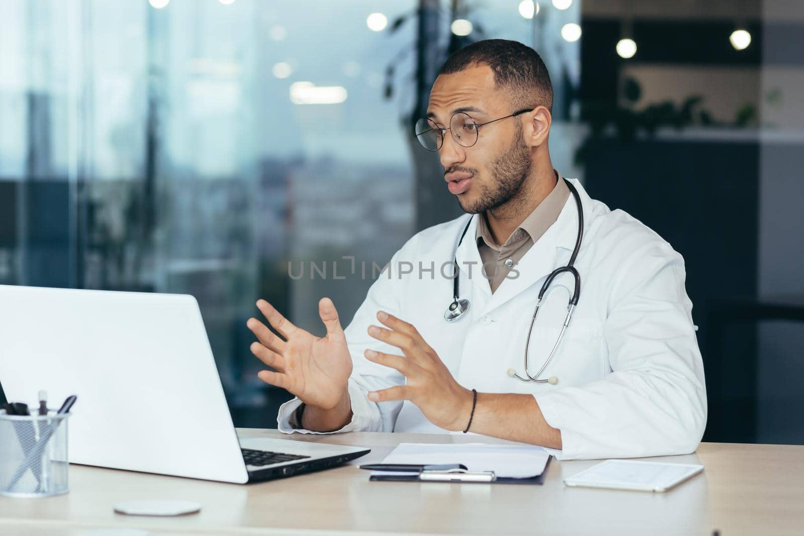 Male family doctor uses laptop for video call and online consultation, serious and concentrated hispanic man in medical coat and stethoscope by voronaman