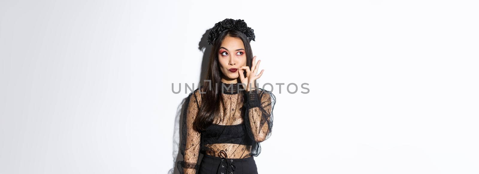 Young gothic woman in black lace dress, dressed-up like witch for halloween party, making seal on lips, zipping mouth as making promise keep secret, standing over white background by Benzoix