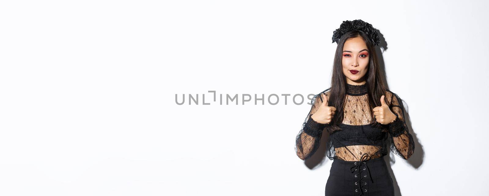Pleased sassy attractive woman in witch dress showing thumbs-up in approval, like and praise your choice, making compliment, standing over white background.