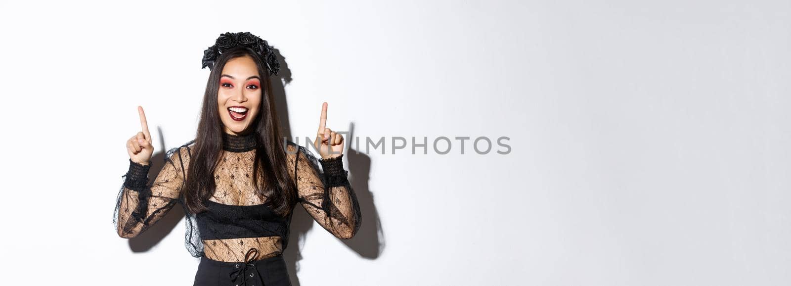 Excited smiling woman celebrating halloween in black gothic dress and wreat. Asian girl wearing witch costume and pointing fingers up, standing white background by Benzoix