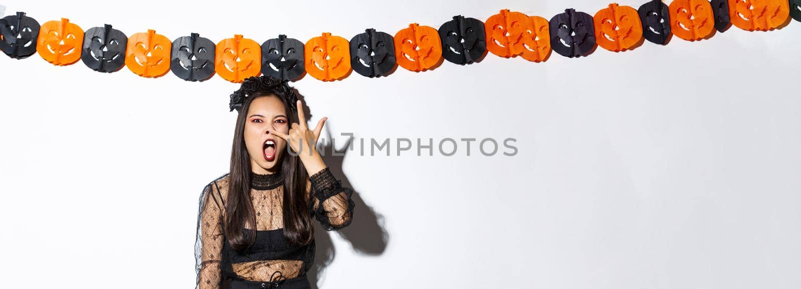 Image of funny asian woman in witch costume grimacing, wearing gothic lace dress and standing against pumpkin streamers, decorations for halloween.