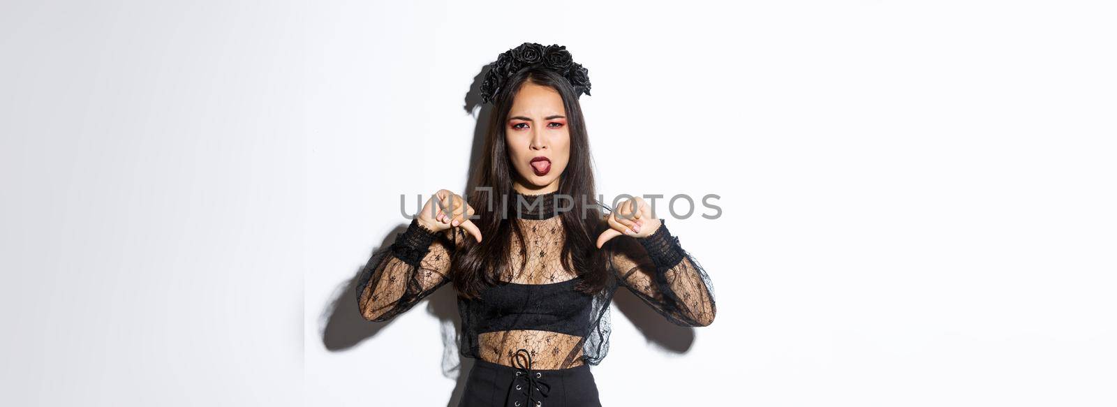 Image of disappointed asian woman wearing halloween costume of witch, showing thumbs-down and sticking tongue displeased, white background.