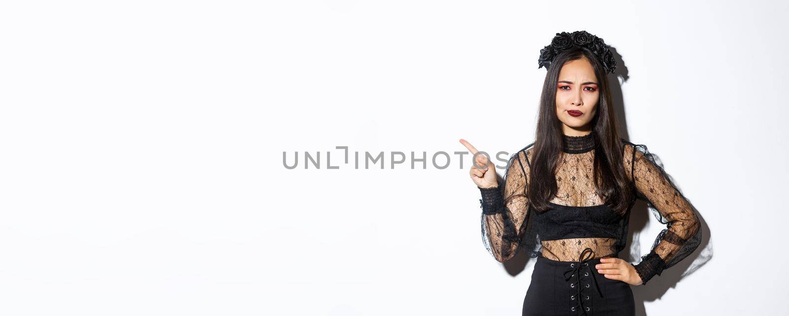 Image of disappointed and skeptical asian woman in witch costume complaining on something, pointing upper left corner and grimacing dissatisfied, standing over white background in halloween dress.