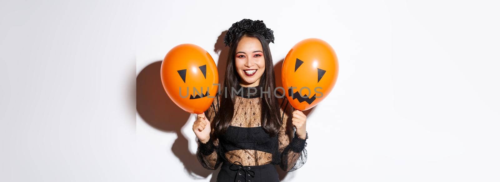 Image of cheerful asian woman in witch costume celebrating halloween, holding balloons with scary faces, standing over white background by Benzoix