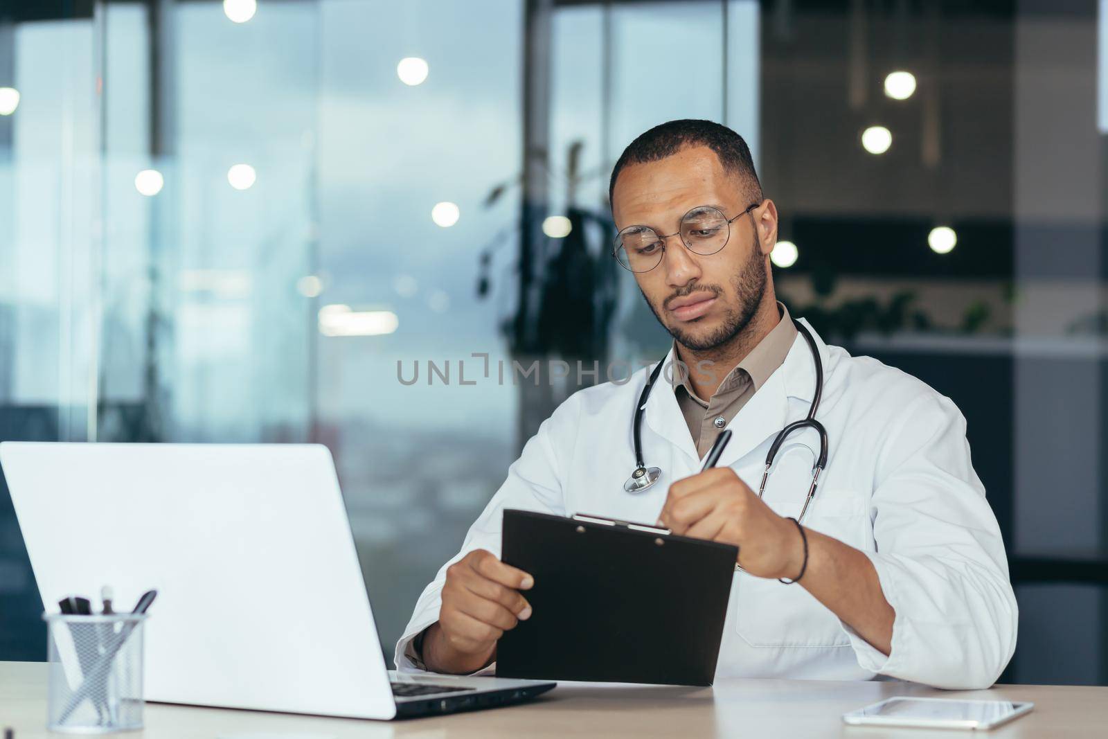 Serious and focused hispanic doctor in medical coat and stethoscope behind paperwork, man in glasses working inside clinic office using laptop.