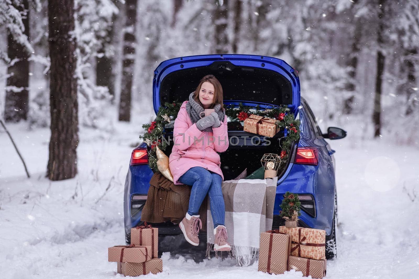 A woman in winter clothes drinks a hot drink sitting in a car decorated in a New Year's way. Traveling by car through the forest. A trip before Christmas.
