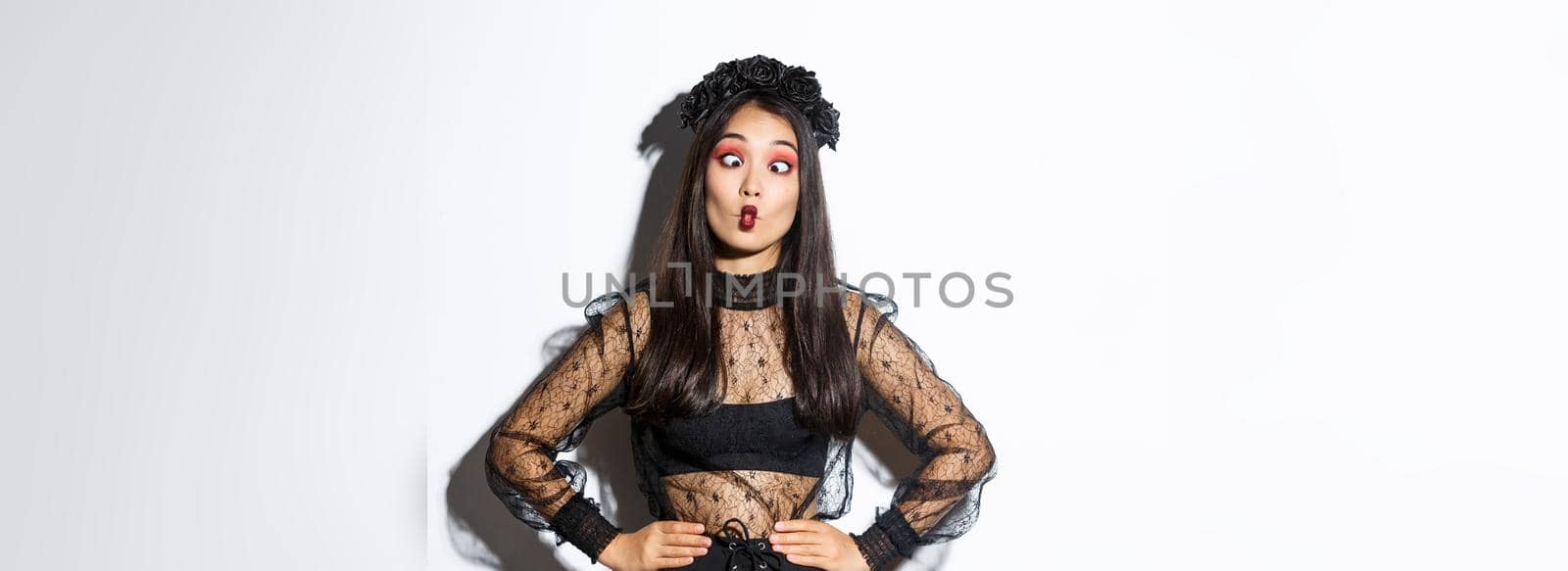 Image of funny asian girl celebrating halloween, wearing gothic lace dress to impersonate witch, showing silly faces and having fun, standing over white background by Benzoix