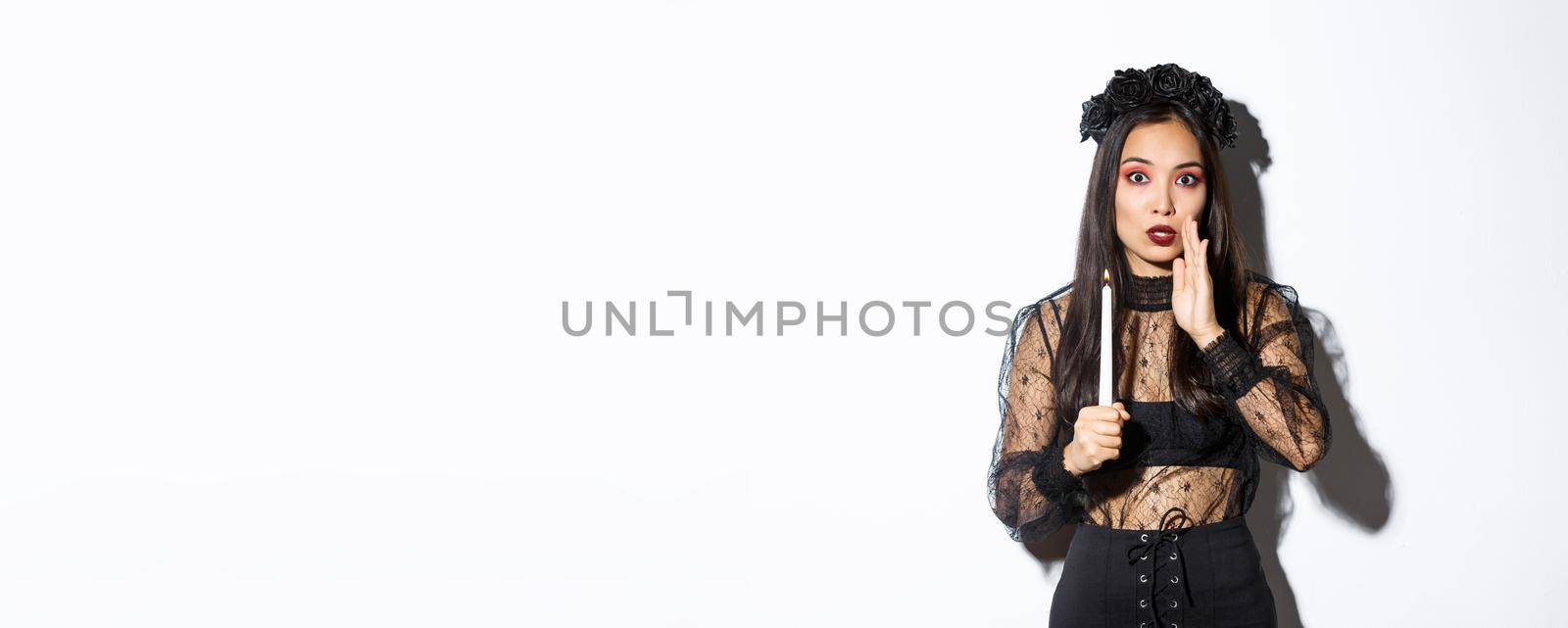 Tensed and worried asian woman in witch costume holding lit candle and whispering, standing over white background.
