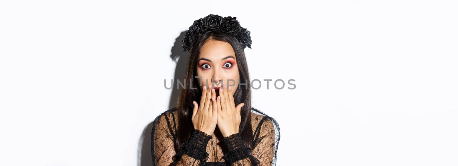 Close-up of shocked asian woman in gothic lace dress and wreath gasping, cover mouth and stare at camera amazed, standing over white background by Benzoix