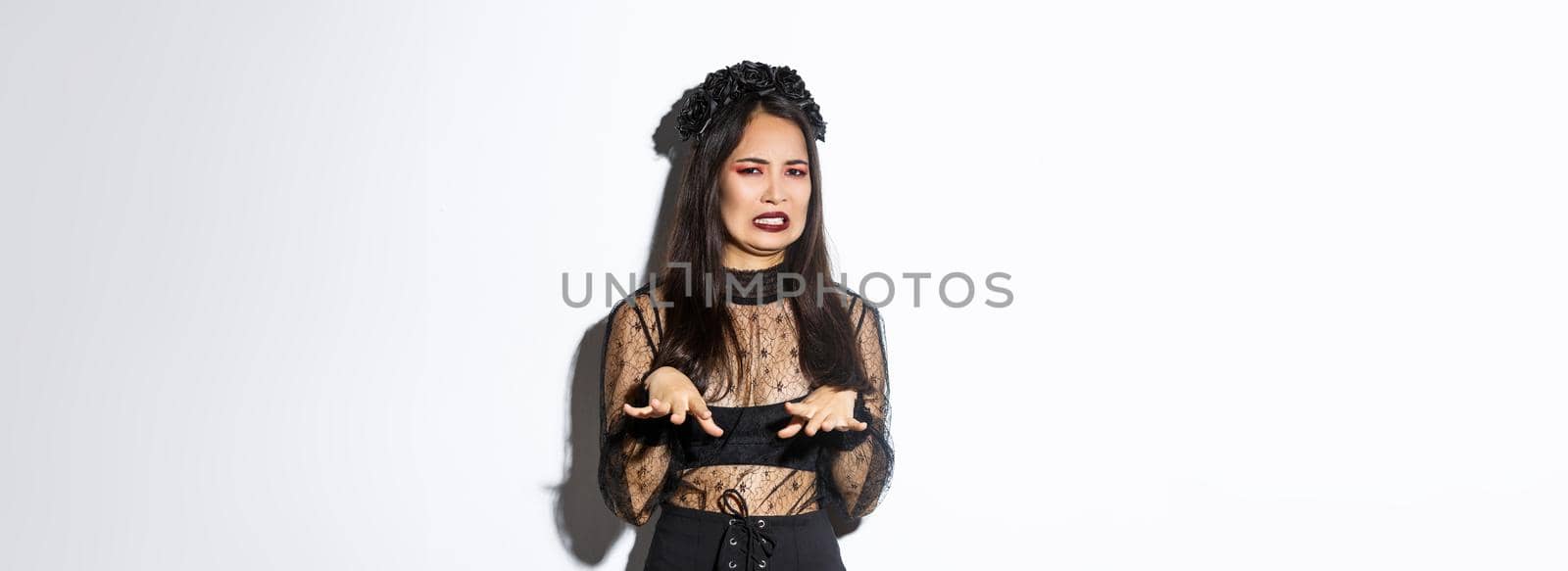 Image of funny asian woman in halloween witch costume looking disgusted, refuse from something nasty or creepy, standing over white background, express aversion.
