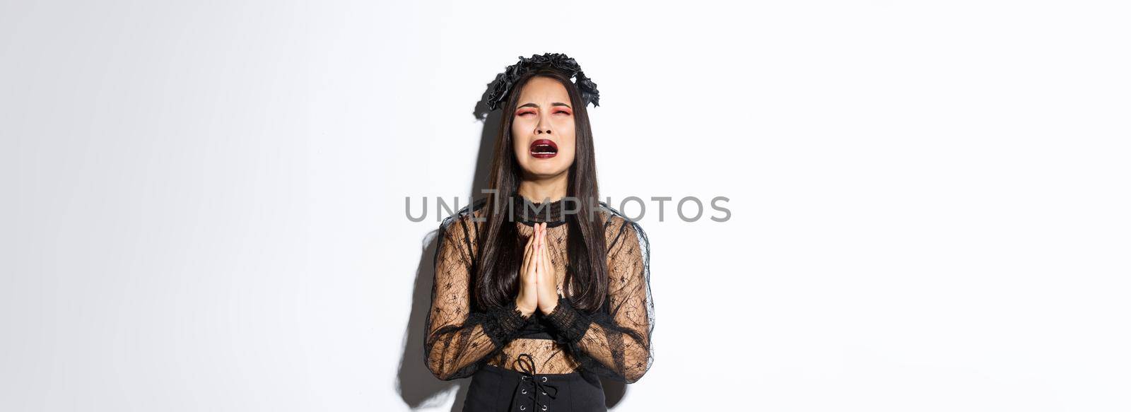 Miserable young asian woman in trouble pleading god, crying and begging for help, wearing halloween gothic dress and wreath, supplicating over white background by Benzoix