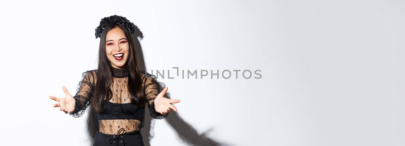 Cheerful smiling beautiful woman in black lace dress and gothic makeup inviting someone. Asian girl spread hands sideways and smiling, welcome come closer, standing white background.